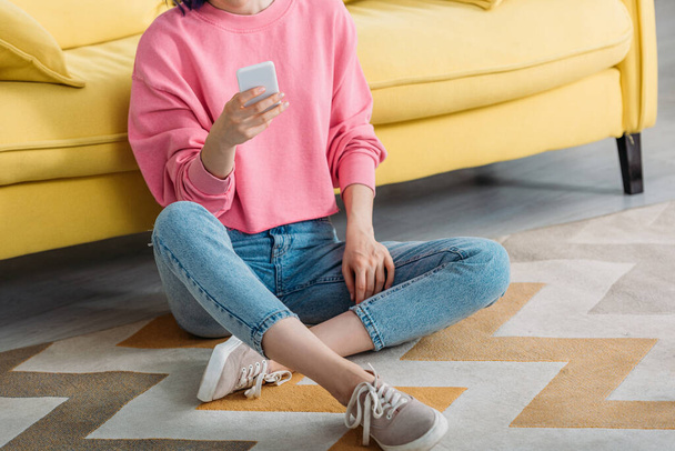 Cropped view of woman with crossed legs holding smartphone near sofa on floor - Foto, afbeelding