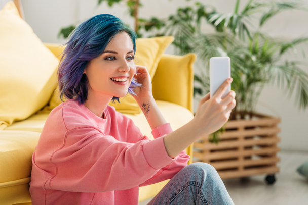 Fille aux cheveux colorés souriant et prenant selfie avec smartphone près du canapé sur le sol dans le salon
 - Photo, image