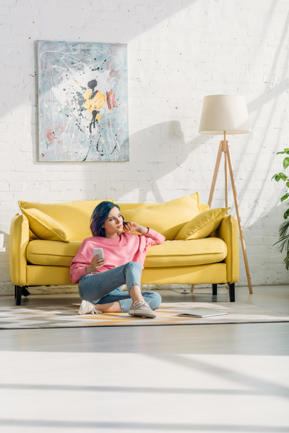 Mulher pensativa com cabelo colorido e smartphone sentado perto do sofá no chão na sala de estar
 - Foto, Imagem