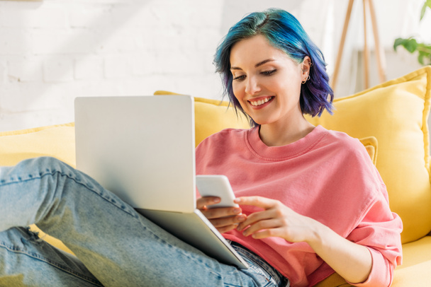Фрилансер с красочными волосами и ноутбуком, держащий смартфон, улыбающийся и лежащий на диване
 - Фото, изображение