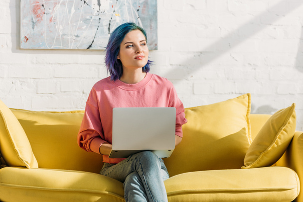 Freelancer with colorful hair and laptop looking away and sitting on yellow sofa with crossed legs in living room - Photo, Image