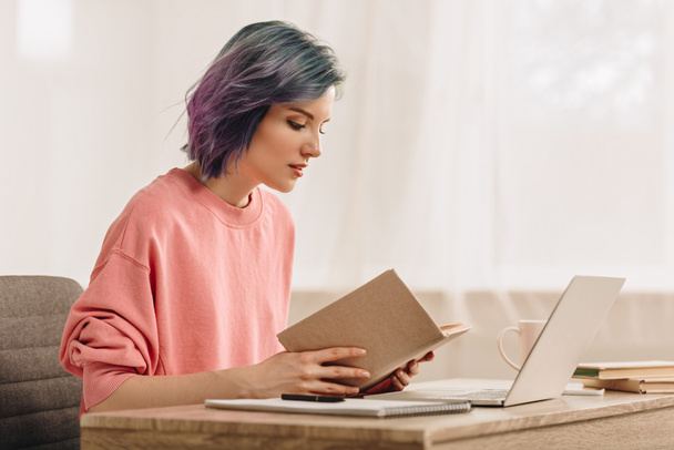 Freelancer with colorful hair reading book near laptop at table in living room - Foto, Bild