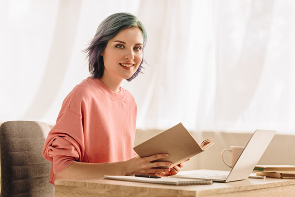 Freelancer with colorful hair and book looking at camera and smiling at table with laptop in living room - Photo, Image