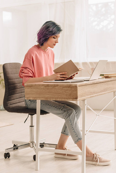 Freelancer με πολύχρωμο βιβλίο ανάγνωσης μαλλιών στο τραπέζι με φορητό υπολογιστή στο σαλόνι - Φωτογραφία, εικόνα
