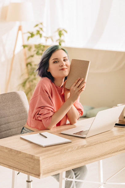 Freiberuflerin mit buntem Haar hält Buch in der Hand, blickt in die Kamera und lächelt neben Laptop und Notizbuch am Wohnzimmertisch - Foto, Bild
