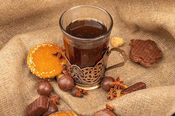 Огранённый стакан чая в винтажном держателе для чашки, печенье, шоколадные торты и шоколад на фоне текстурированной ткани. Закрыть
 - Фото, изображение
