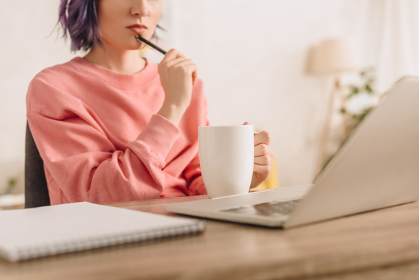 Freiberufler mit buntem Haar hält Tasse Tee und Stift am Tisch mit Notizbuch und Laptop im Wohnzimmer - Foto, Bild