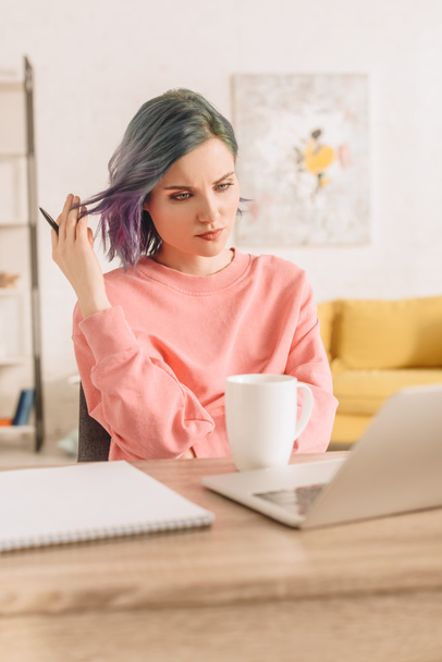 Enfoque selectivo de freelancer reflexivo con lápiz de retención de pelo colorido en la mesa con computadora portátil, taza de té y portátil en la sala de estar
 - Foto, Imagen