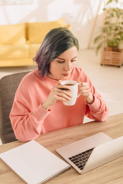 High angle view of freelancer with colorful hair holding cup of tea at table with laptop and notebook in living room - Foto, Imagen