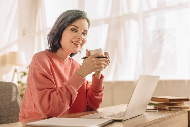 Freelancer com cabelo colorido, xícara de chá e caneta olhando para a câmera e sorrindo à mesa com laptop na sala de estar
 - Foto, Imagem