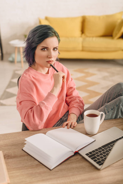 Hoge hoek uitzicht van freelancer met kleurrijk haar en pen kijken naar de camera aan tafel met kopje thee, notebook en laptop in de woonkamer - Foto, afbeelding