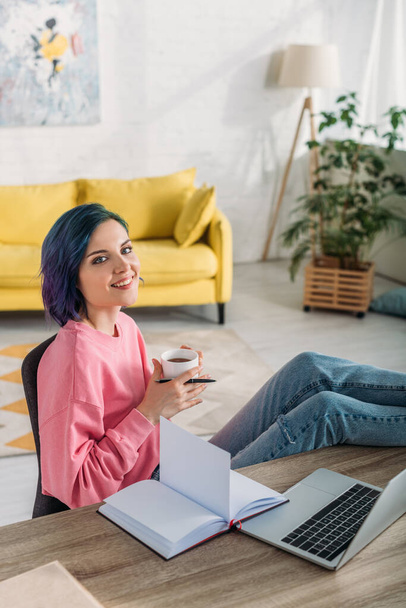 Hochwinkelblick auf Freelancer mit buntem Haar, Tasse Tee und Stift lächelnd und Beine auf den Wohnzimmertisch legen - Foto, Bild