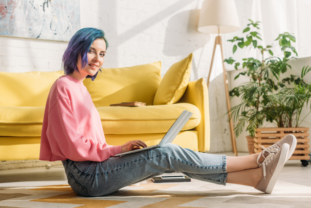 Freelancer con el pelo colorido sonriendo, mirando a la cámara y trabajando con el ordenador portátil en el suelo en la sala de estar
 - Foto, imagen
