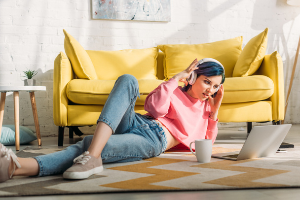Szabadúszó színes haj és fejhallgató nézi laptop és fekvő közel kanapé és csésze tea a nappaliban - Fotó, kép