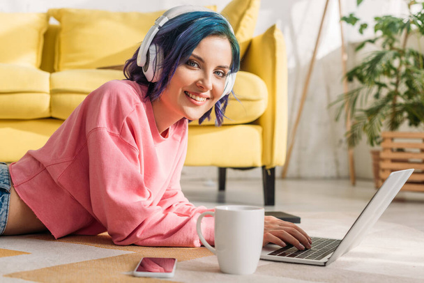 Freelancer com cabelo colorido em fones de ouvido trabalhando com laptop perto de xícara de chá e smartphone, sorrindo e olhando para a câmera no chão
 - Foto, Imagem