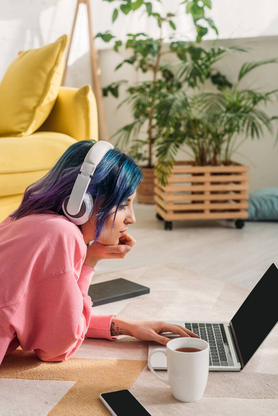 Freelancer met kleurrijk haar en koptelefoon werken met laptop in de buurt van kopje thee, notebook en smartphone op de vloer - Foto, afbeelding