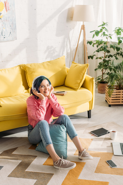 High angle view of freelancer with colorful hair in headphones smiling near sofa on floor in living room - Foto, Imagen