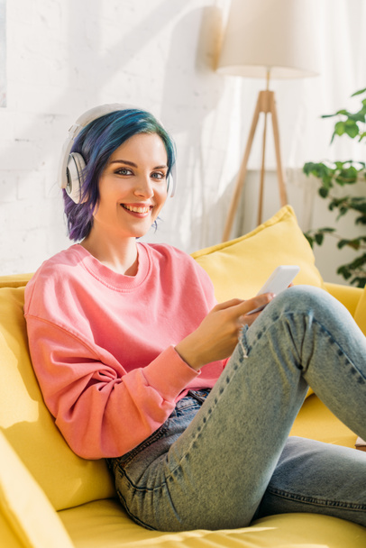 Kobieta z kolorowymi włosami i słuchawkami trzymając smartfona, uśmiechając się i patrząc na aparat na kanapie - Zdjęcie, obraz