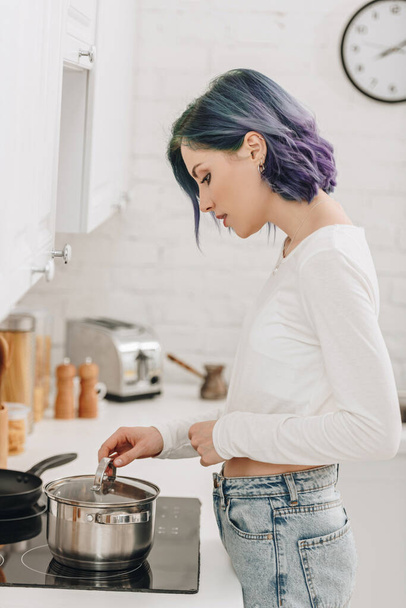 Girl with colorful hair preparing food and touching pan lid near kitchen stove - Фото, зображення