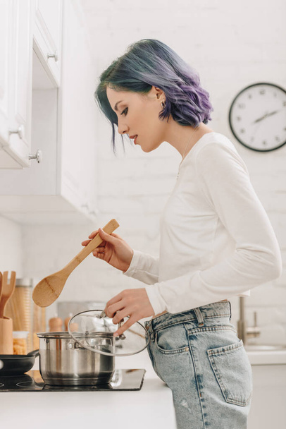 Girl with colorful hair holding pan lid and spatula near kitchen stove - Photo, Image