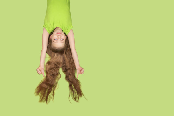 Lustiges und hübsches kleines Mädchen mit extrem langen Haaren, das isoliert auf grünem Hintergrund lächelt. Mädchen in grünem T-Shirt, kopfüber hängend mit fliegendem Haar. Kopierraum. - Foto, Bild
