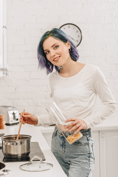 Селективный фокус девушки с красочными волосами, улыбающейся, смотрящей в камеру и готовящей суп на кухне
  - Фото, изображение