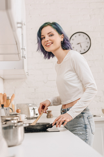 Vista de ángulo bajo de la chica con el pelo colorido preparando la comida con espátula en la sartén, sonriendo y mirando a la cámara en la cocina
 - Foto, Imagen