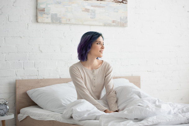 Atrakcyjna kobieta z kolorowymi włosami odwracając wzrok i uśmiechając się na łóżku w sypialni - Zdjęcie, obraz