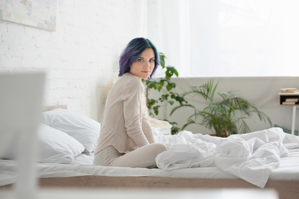 Selectieve focus van de vrouw met kleurrijk haar kijken naar de camera en zitten op bed in de slaapkamer - Foto, afbeelding