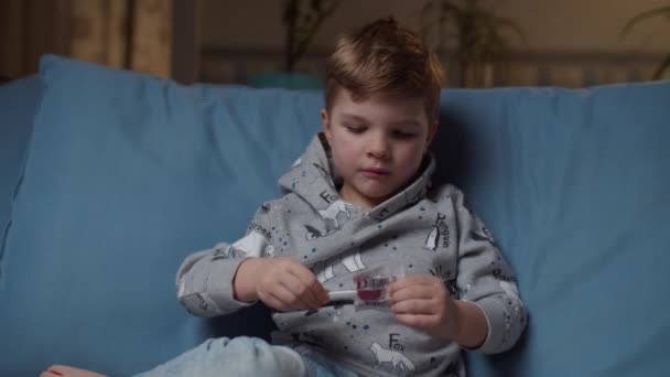 Young boy opening sweets and take lollipop to the mouth sitting on cozy couch at evening night. Kid enjoying hard candy and smiling looking at camera.  - Materiał filmowy, wideo