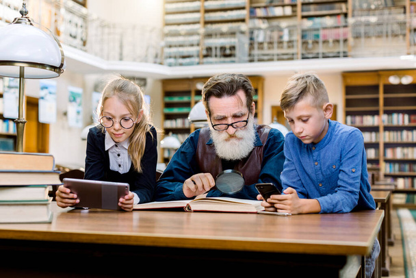 Family, education, digital technologies vs book. Elderly man reading book, while his smart teen students or grandchildren using modern gadgets, girl holds ebook or tablet, boy holds smartphone - Photo, Image
