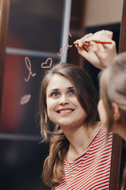 reflection of young woman writing with cosmetic pencil words "I love you" in mirror, portrait of romantic cute girl, concept declaration of love - Photo, Image