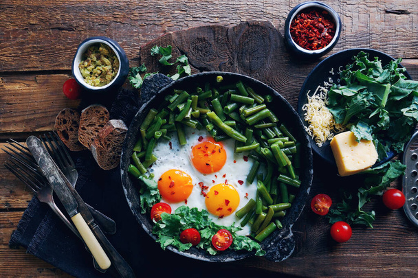 Pan of fresh fried eggs with tomatoes, green beans, bread, spices and herbs on rustic wooden background, healthy breakfast concept, top view - Photo, image