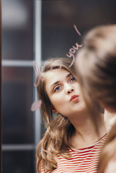 reflection of young woman face in mirror with inscription "I love you", painted heart and lip kiss, happy girl in romantic relationship, concept creative declaration of love - Фото, изображение