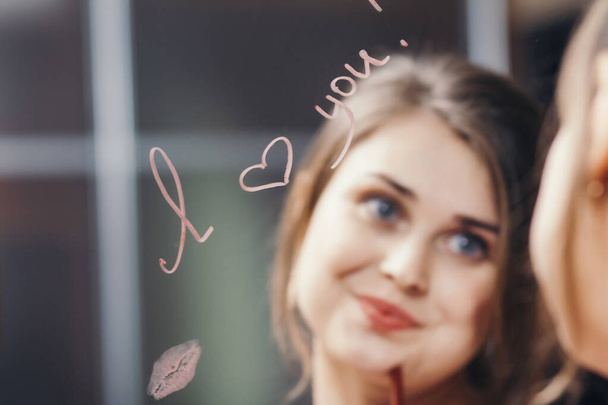 reflection of young woman face in mirror with inscription "I love you", painted heart and lip kiss, happy girl in romantic relationship, concept creative declaration of love - Foto, afbeelding