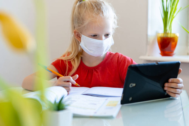 Distance learning online education. Sickness schoolgirl in medical mask studying at home with digital tablet in hand and doing school homework. Training books and notebooks on table. - Foto, imagen