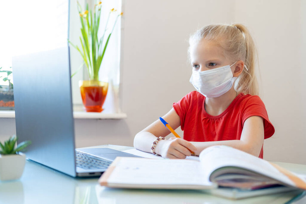 Distance learning online education. Sickness schoolgirl in medical mask studying at home, looking at laptop notebook and doing school homework. Training books and notebooks on table. - 写真・画像