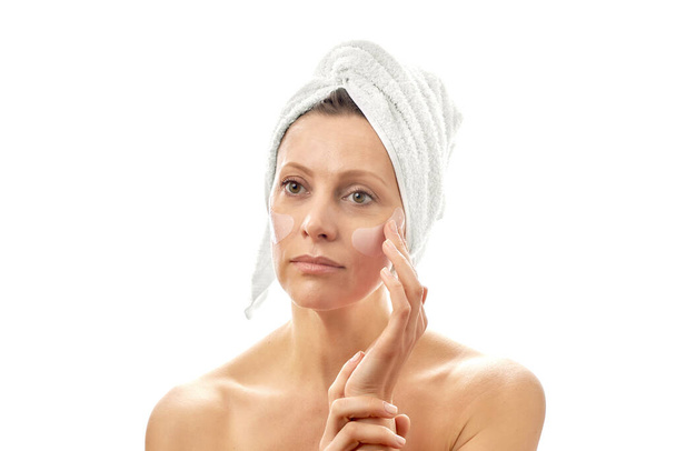 Kafasında beyaz bir havlu olan güzel bir kadın göz bantlarıyla yüz yenileme ameliyatı yapıyor. Beyaz izole arkaplan - Fotoğraf, Görsel