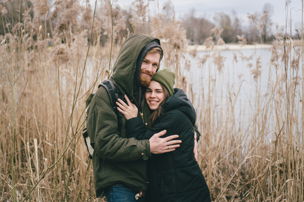 Mladý zamilovaný bělošský pár stojí v objetí u jezera a rákosí. Víkendy a procházky v přírodě. Venkovní rekreace v zemi. Aktivní dovolená a turistika na podzim. - Fotografie, Obrázek