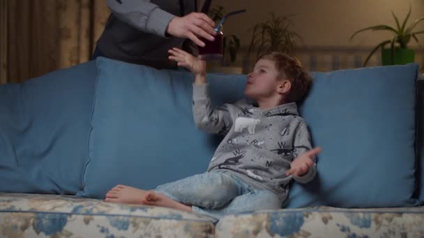 Blond preschooler boy drinking juice sitting on cozy couch at evening. Mother brings son glass of drink with straw. Kid drinking blueberry juice with non-plastic straw on sofa at home.  - Кадри, відео