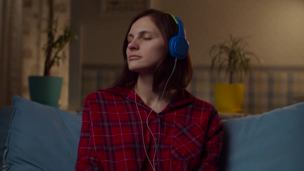 Młoda kobieta po trzydziestce w czerwonej koszuli słuchająca muzyki w kolorowych słuchawkach siedząca na kanapie w domu. Kobieta ciesząca się muzyką z zamkniętymi oczami w zwolnionym tempie.  - Materiał filmowy, wideo