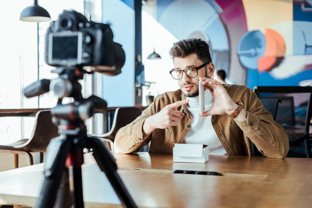 Foco seletivo do blogueiro na frente da câmera digital apontando no smartphone à mesa no espaço de coworking
 - Foto, Imagem