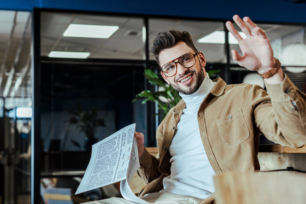 Selective focus of IT worker with newspaper waving hand and smiling in coworking space - Photo, Image