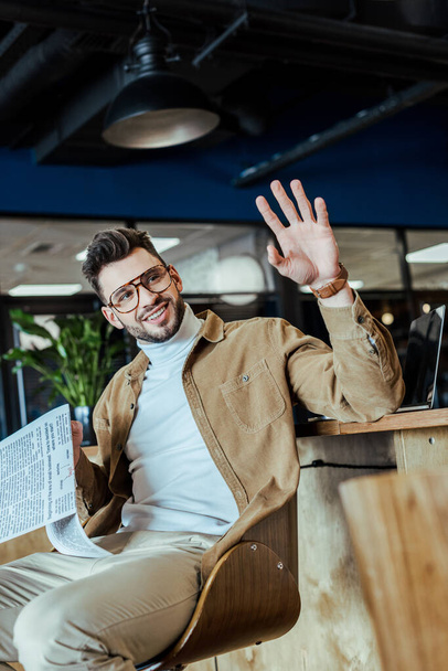 Selective focus of IT worker holding newspaper, waving hand and smiling in coworking space - Zdjęcie, obraz