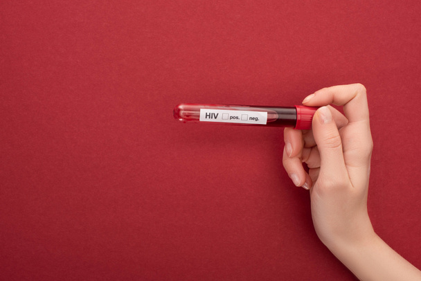 Kırpılmış görüşlü kadın, kırmızı arka planda HIV harfleriyle test tüpünde kan örneği tutuyor. - Fotoğraf, Görsel