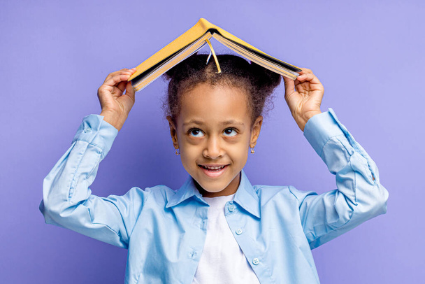 portrait of little mulatto child thinking and holding book on head - Фото, изображение