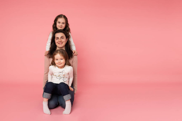 Beautiful young mother with her two daughters sitting on the ground one by one. Looking directly at the camera. Isolated on the pink background - Foto, immagini