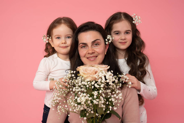 Крупним планом фото здорової красивої сім'ї, молода мама тримає букет весняних квітів, її маленькі дочки з квітами волосся, що стоять поруч і дивляться на камеру
 - Фото, зображення