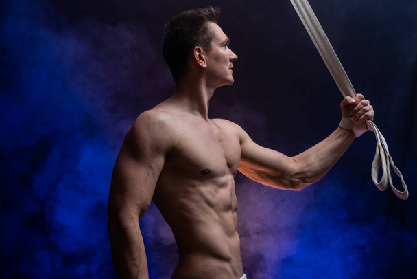Muscular male circus artist with aerial straps on black and smoked background - Foto, imagen