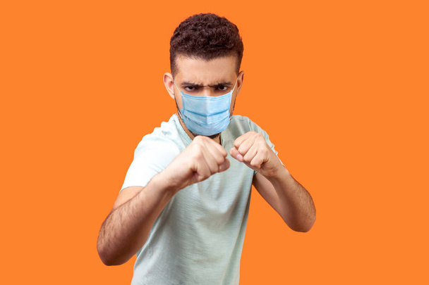 Boxing and self-defence. Portrait of weak angry man with medical mask in casual white t-shirt frowning and keeping fists clenched, ready to defend in fight. studio shot isolated on orange background - Photo, image